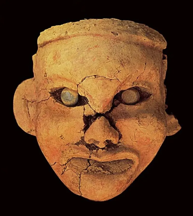 Museum reference photo: the clay goddess head from the Hongshan Goddess Temple at Niuheliang — a cracked earthen face with jade-inlaid eyes and a faint smile