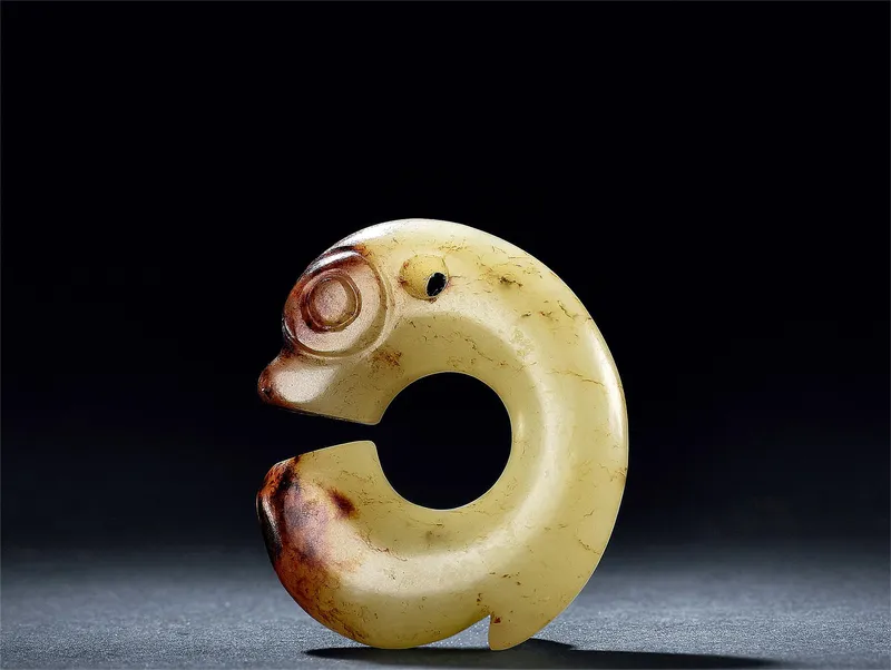 Museum reference photo: a Hongshan jade pig-dragon (zhulong) — pale golden jade with russet staining, curled head-to-tail into a C-shaped ring
