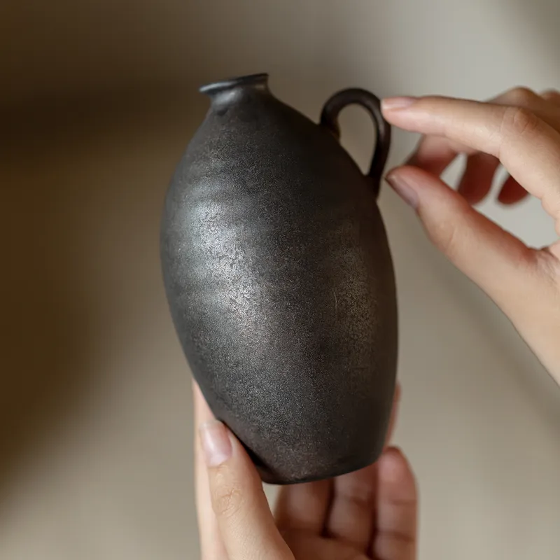 Concept visualization: a matte black hand-finished stoneware pourer, cradled in two hands — an atmosphere study for the Spiral Pourer
